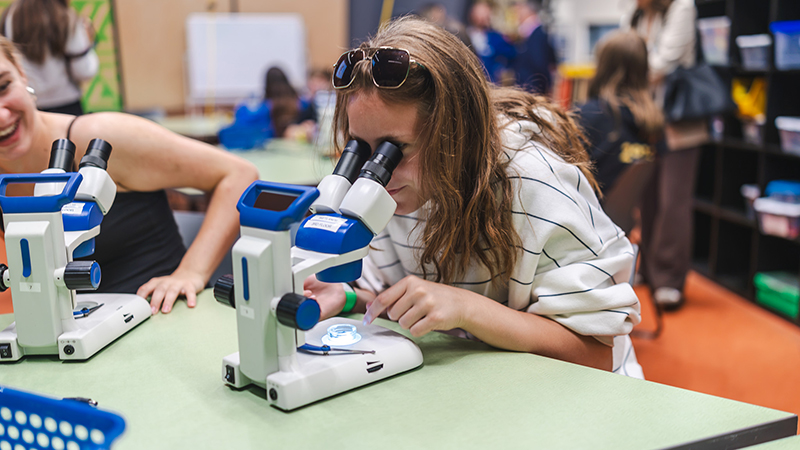 Élève utilisant un microscope pour observer un échantillon dans le cadre d’une activité scientifique pratique, tandis que d’autres participantes et participants travaillent à des tables en arrière-plan.